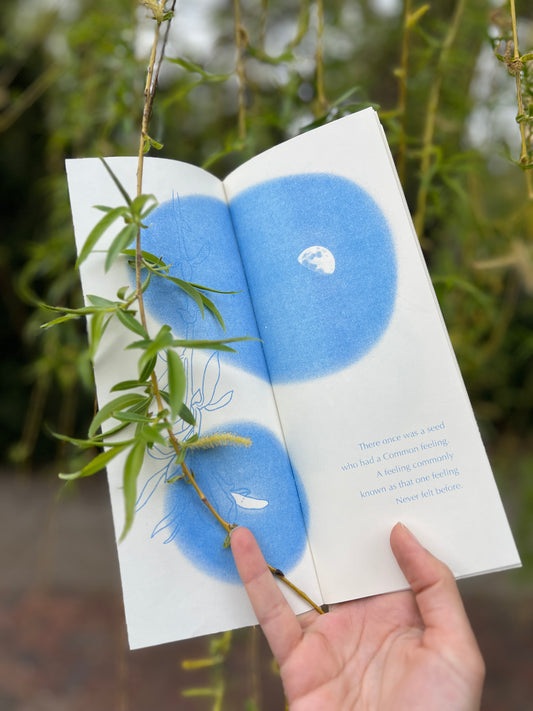 A photograph of Jo’s hand holding open the first spread of the book, their pinky finger gently pinning a single willow branch to the paper. The live branch emulates the illustration below, a line drawing of a similar willow tendril with a small seed looking up to a gibbous moon in a night sky. 