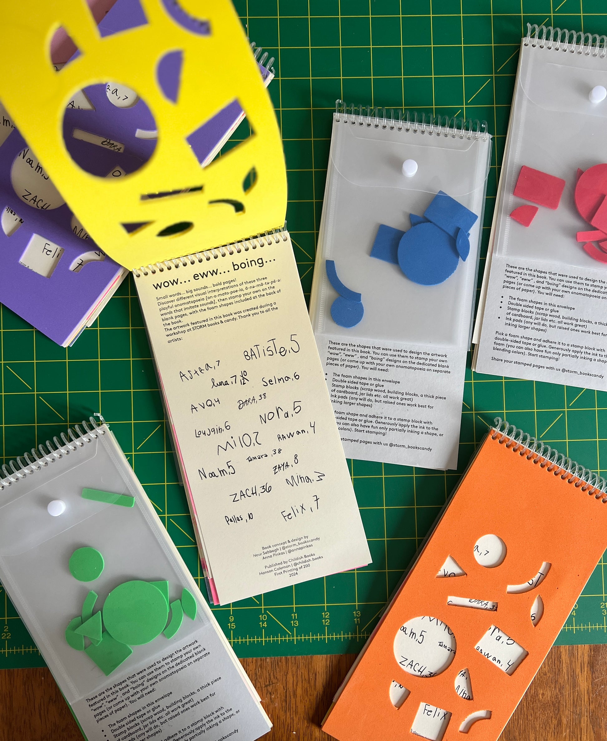 a mess of six different copies of the book playfully sprawl out over a work table with a large green cutting mat. One is being peeled open to the title page, while others lay either on their faces or backs. 