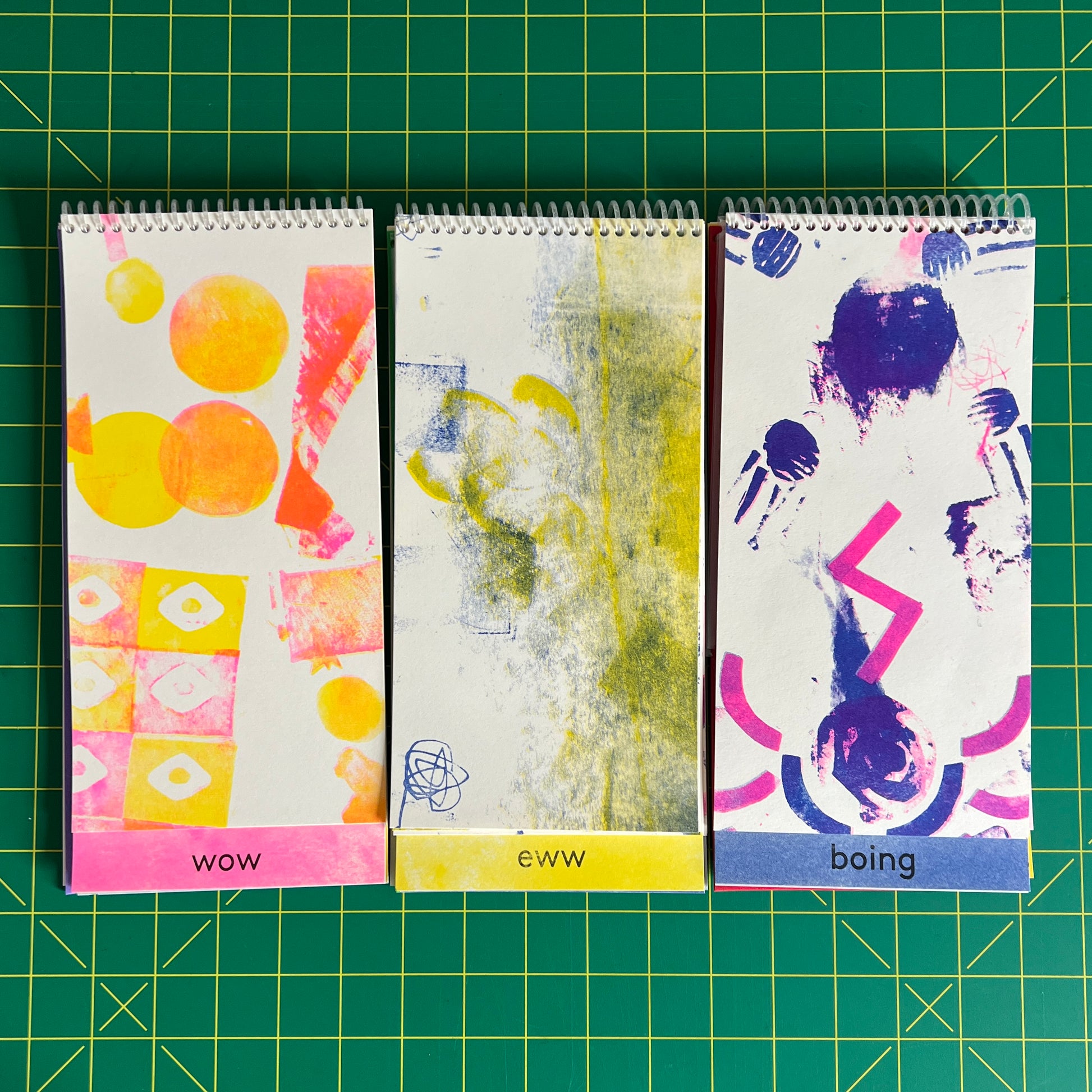 Three copies of the book are open to different pages from each of the three sections "wow", "ew", and "boing". Each section of artwork has been risograph printed in different two tone colorways: Wow in fluro pink and yellow inks, ew in yellow and medium blue, and boing in fluro pink and medium blue!